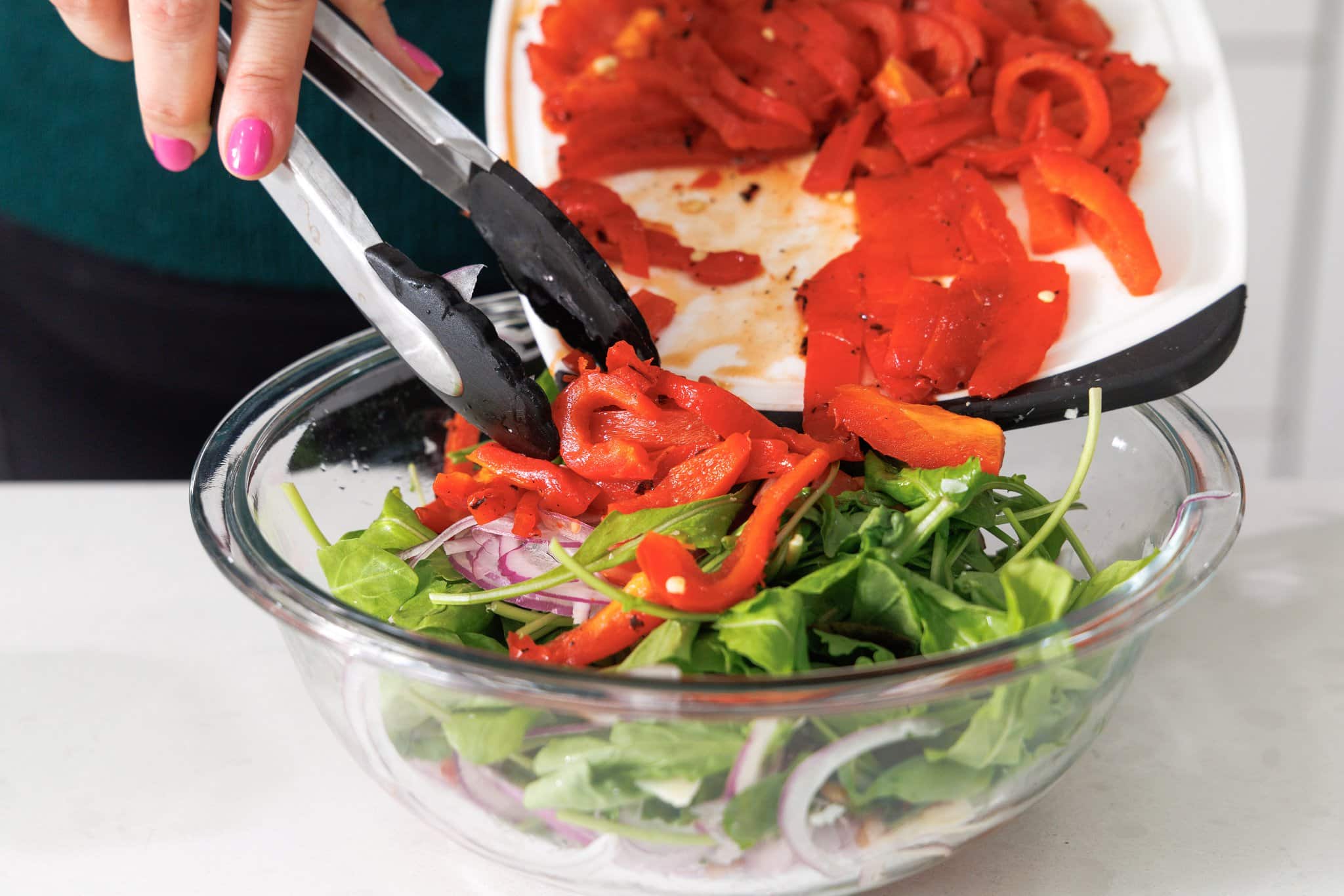 Adding roasted peppers to a salad in a glass bowl.